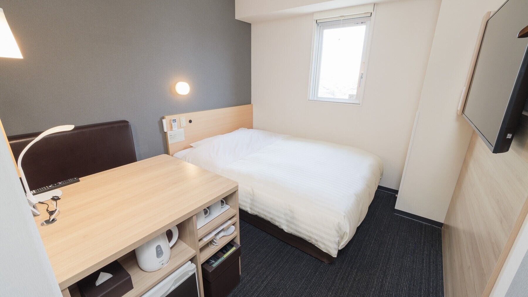 Une chambre d’hôtel compacte comprenant un lit simple, un bureau avec une chaise, une petite table avec une bouilloire, un téléviseur fixé au mur et une fenêtre laissant entrer la lumière naturelle.