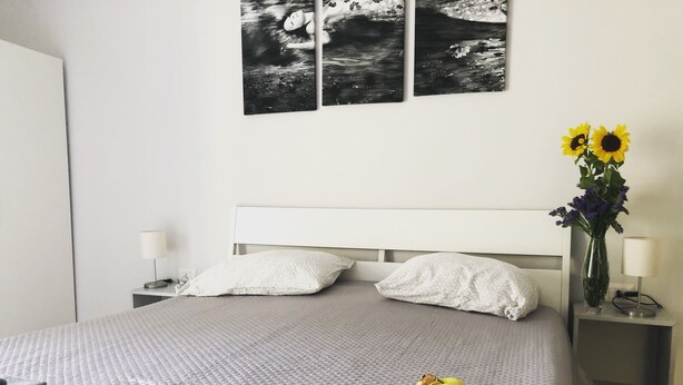 En snyggt bäddad säng med ett grått täcke, två kuddar, ett sängbord med en lampa, en vas med blommor och en fruktskål på sängen.