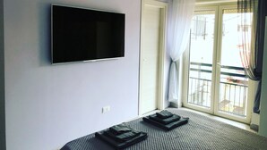 Ett rum med en platt-TV som är monterad på väggen, en säng med ett mönstrat överkast och utsikt över en balkong med ett räcke.
