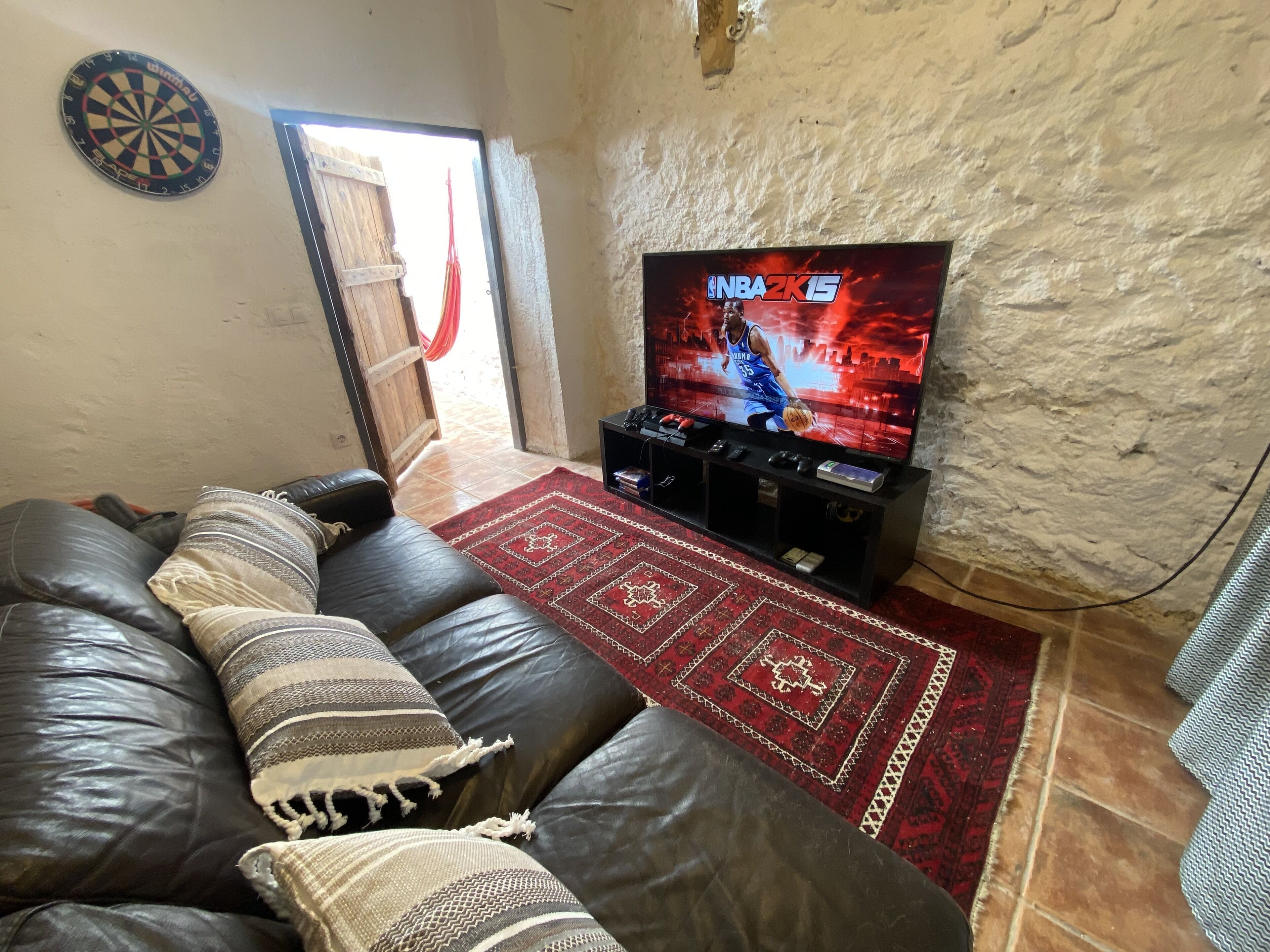 A living room with a leather sofa, a red patterned rug, a TV displaying NBA 2K15, and a dartboard on the wall.
