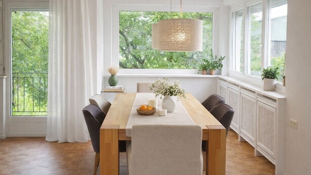 A dining area with a wooden table, chairs, a hanging light fixture, and a view of greenery through large windows.