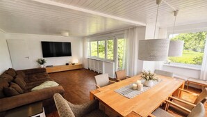 A modern living room with a wooden dining table, a sofa, and a TV mounted on the wall.