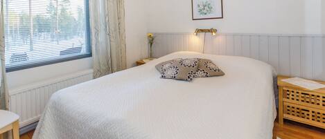 Een slaapkamer met een groot bed, een nachtkastje, een raam met gordijnen, een wandlamp en een lijst met een afbeelding.