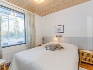 Ett sovrum med en stor säng, ett sängbord, en liten avlastningsyta, ett fönster med gardiner, en vägglampa och en inramad tavla.