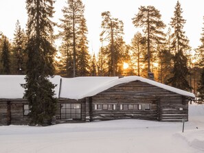 Una cabana de fusta amb un teulat cobert de neu, envoltada d'alts pins i un cel serè.