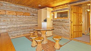 Het interieur van een blokhut, met een houten plafond, een centrale tafel met krukken en een deur aan de rechterkant.