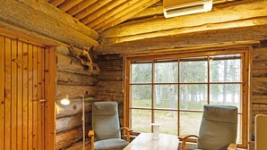 A log cabin interior with a wooden table and four chairs, a large window with a forest view, and a bear head mounted on the wall.