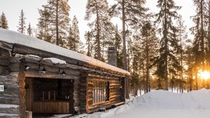 A log cabin with a wooden door and windows, surrounded by snow and tall pine trees.