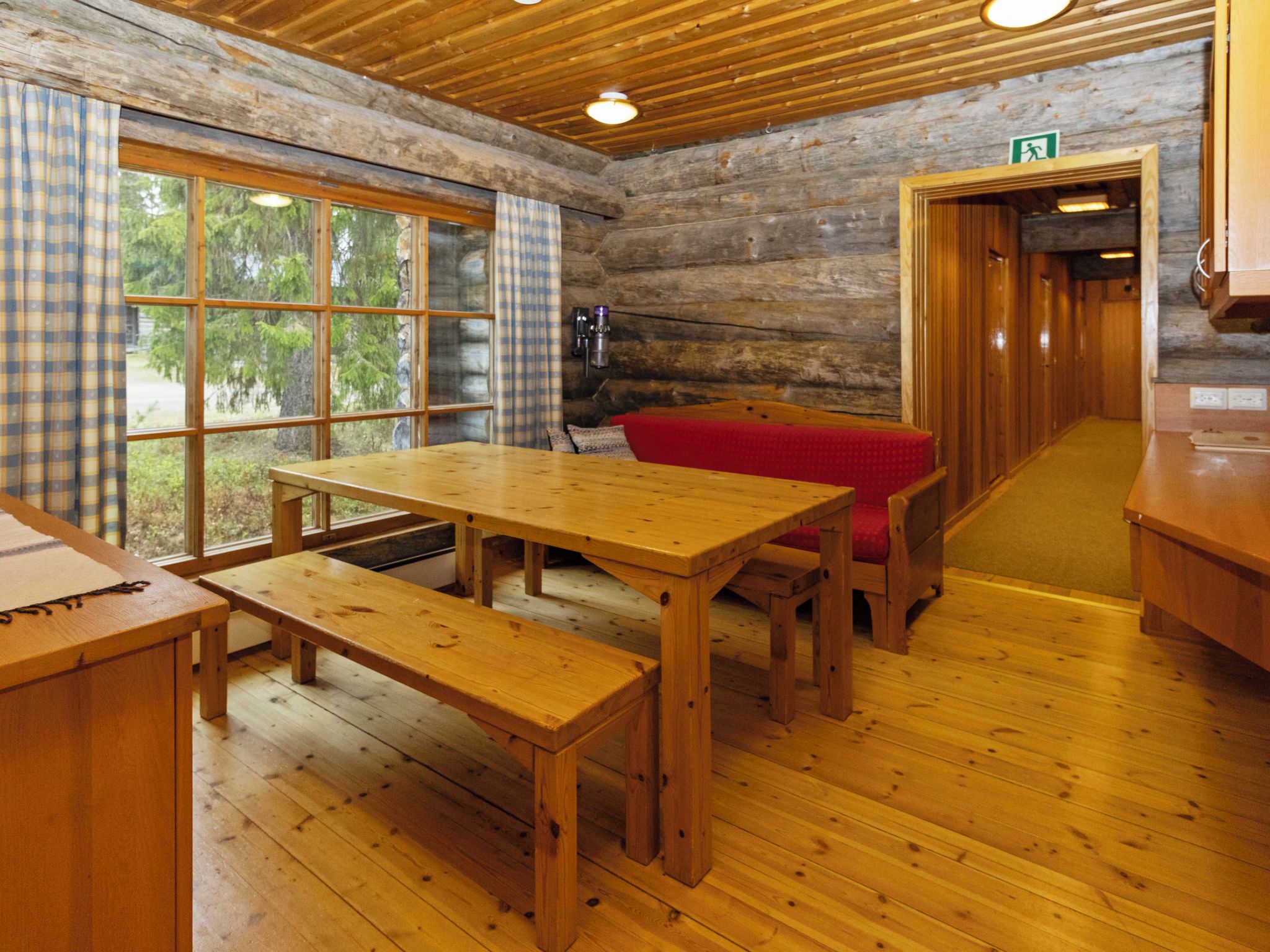 A wooden interior with a wooden table and bench, a red cushioned bench, and a view through large windows.