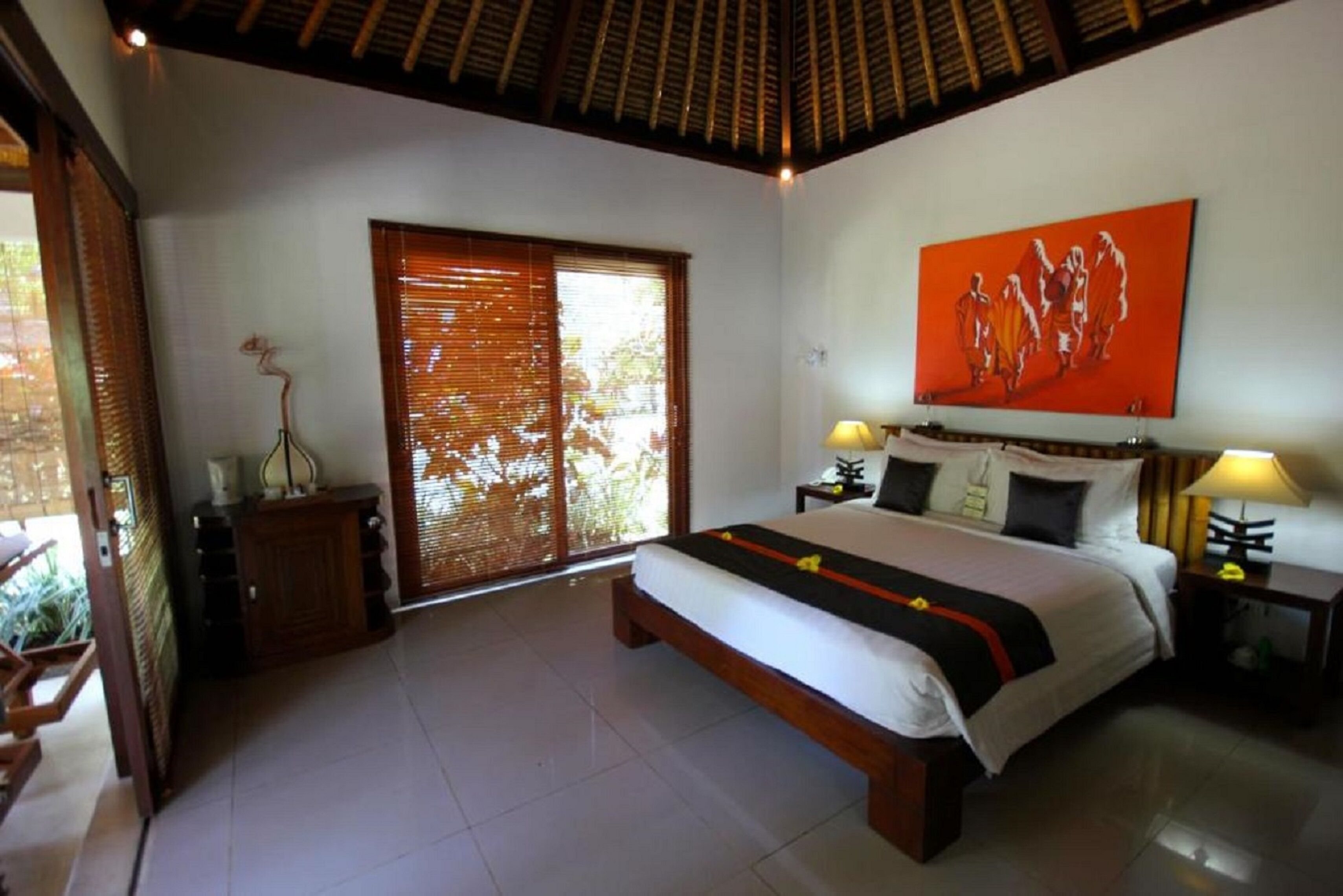 A bedroom with a large bed, a wooden headboard, a painting on the wall, and a window with blinds.