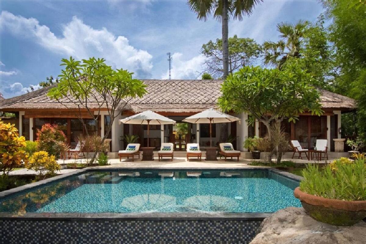 A swimming pool with lounge chairs and umbrellas, surrounded by tropical plants and a building with a tiled roof.