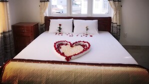 A neatly made bed with a heart-shaped flower arrangement on the bedspread, flanked by two pillows, in a room with a window and curtains.