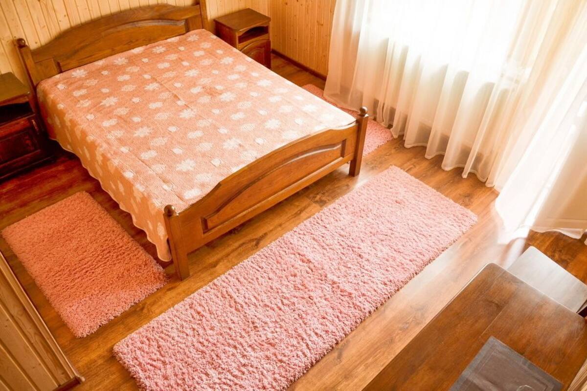 A wooden bed with a patterned bedspread, two pink rugs, a wooden nightstand, and a window with sheer curtains.