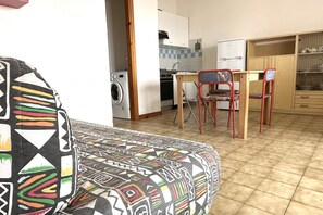 A living room with a patterned sofa, a dining area with a table and chairs, a kitchen with white cabinets and a refrigerator. - M079 - Marcelli, seafront three-room apartment in a residence with swimming pool (Marcelli)