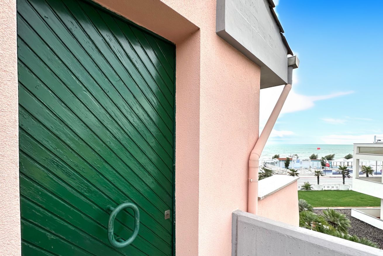 A green door with a metal ring handle, a balcony with a view of a beach and buildings, and a clear sky.