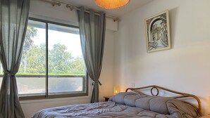 Un dormitorio con cama, ventana con cortinas, mesita de noche con lámpara y un cuadro colgado en la pared.