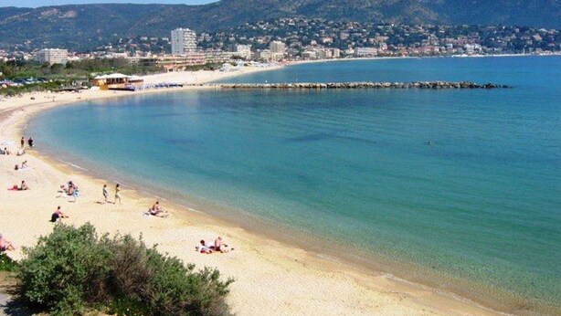 Una playa con aguas cristalinas, una orilla arenosa y edificios y colinas al fondo.