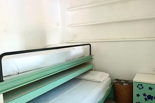 Un lit superposé avec des barreaux métalliques verts, un matelas blanc et un petit tabouret en bois.