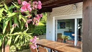 Un tavolo e sedie in legno sotto una pergola fiorita di rosa, una porta finestra e un armadio blu sullo sfondo.
