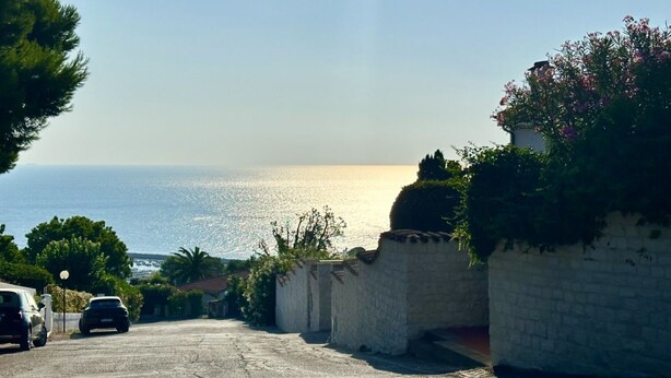 Una strada costiera con auto parcheggiate, cielo sereno e il mare sullo sfondo.