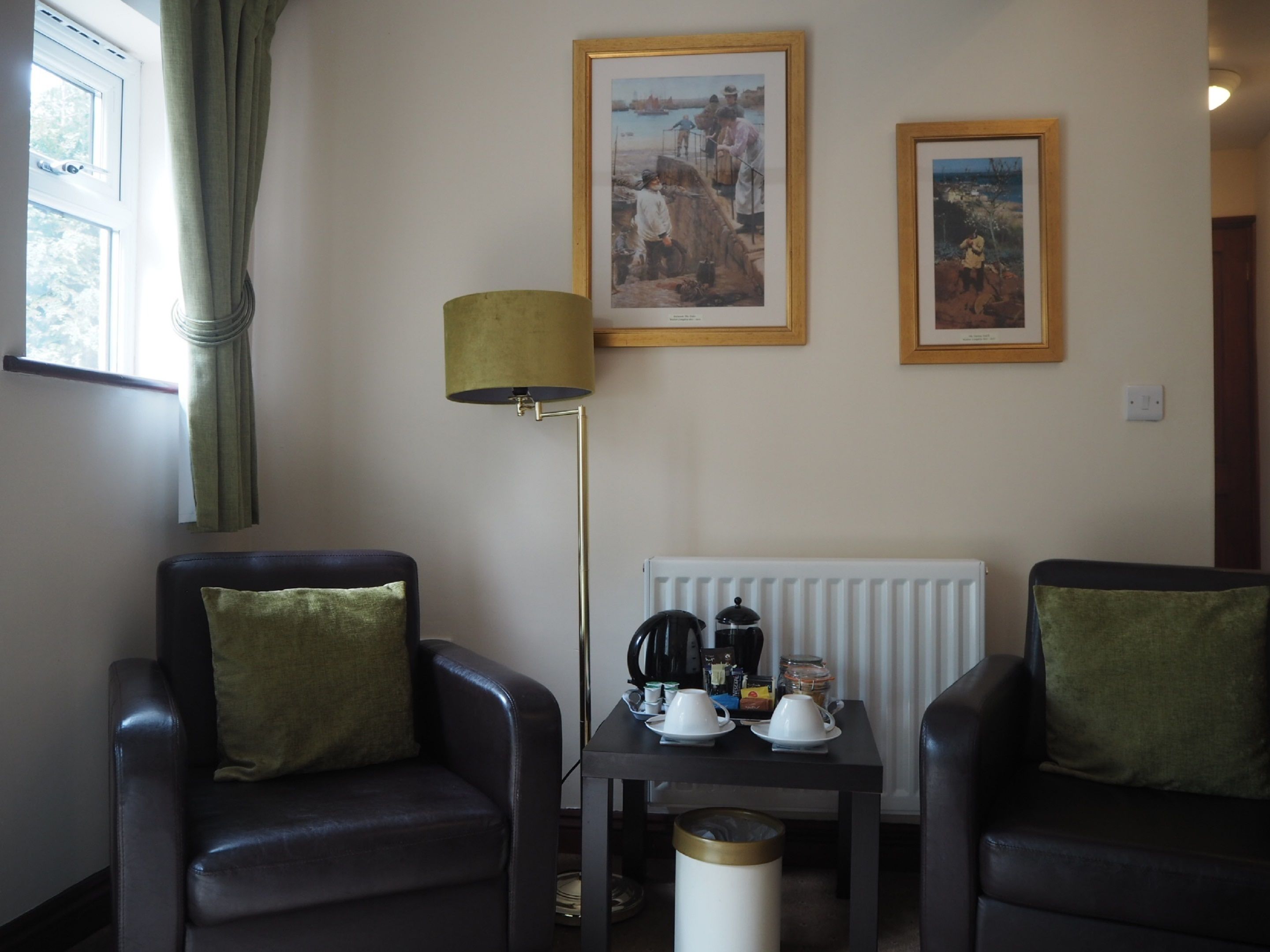 A room with two leather armchairs, a floor lamp, a table with a kettle and cups, and two framed pictures on the wall.