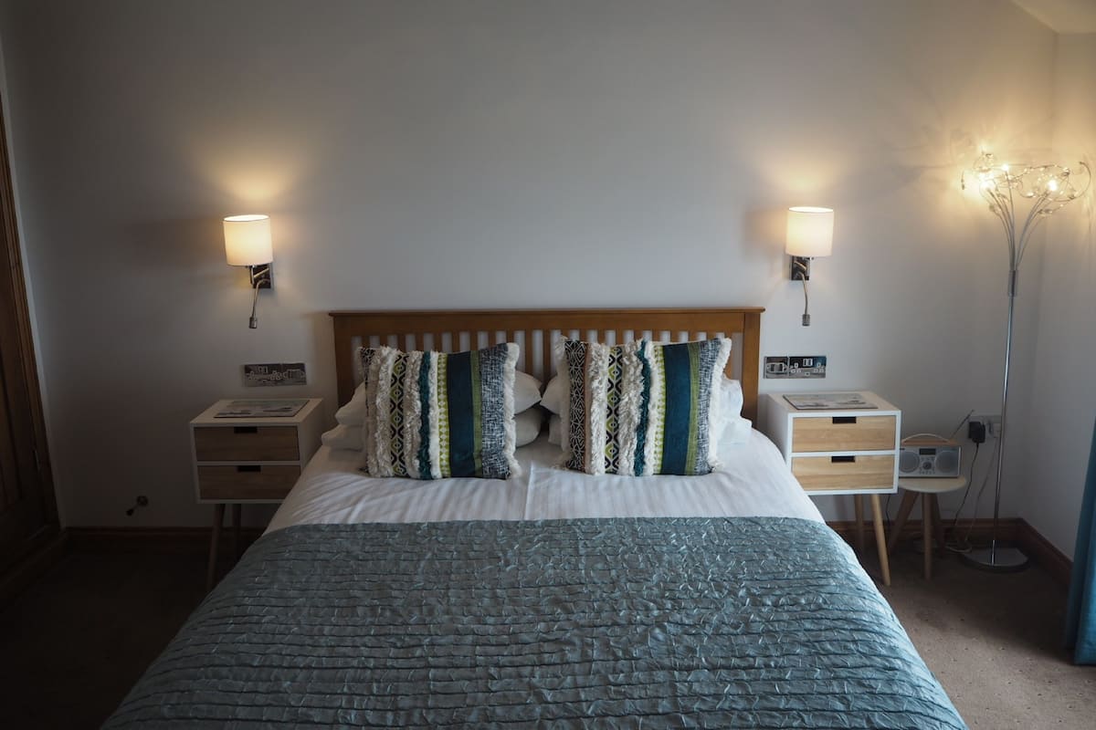 A neatly made bed with a patterned bedspread and multiple pillows, flanked by bedside tables with lamps and a small speaker.