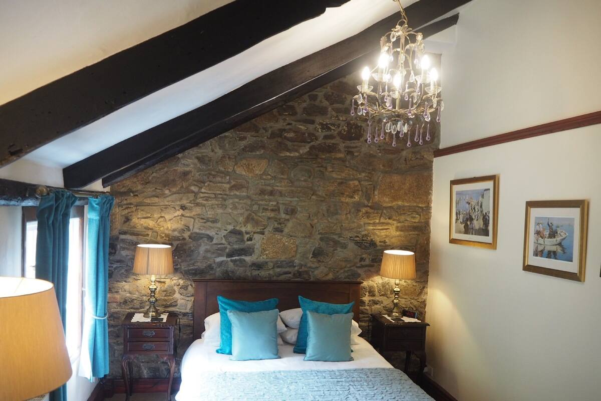 A bedroom with a stone wall, a chandelier, a bed with blue bedding, two bedside tables with lamps, and framed pictures on the wall.