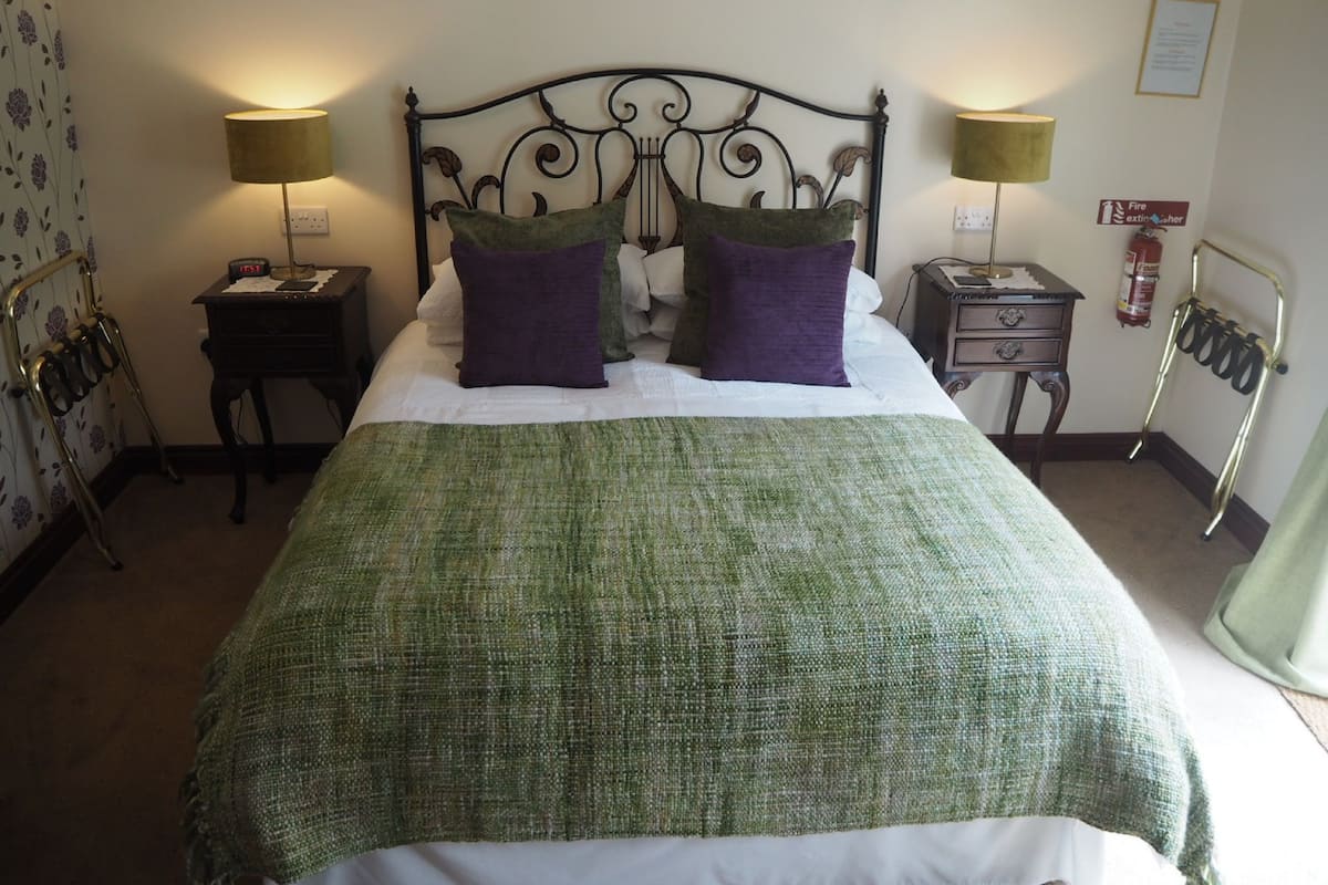 A neatly made bed with a green blanket and two purple pillows, flanked by two bedside tables with lamps, a fire extinguisher, and a metal rack on the wall.