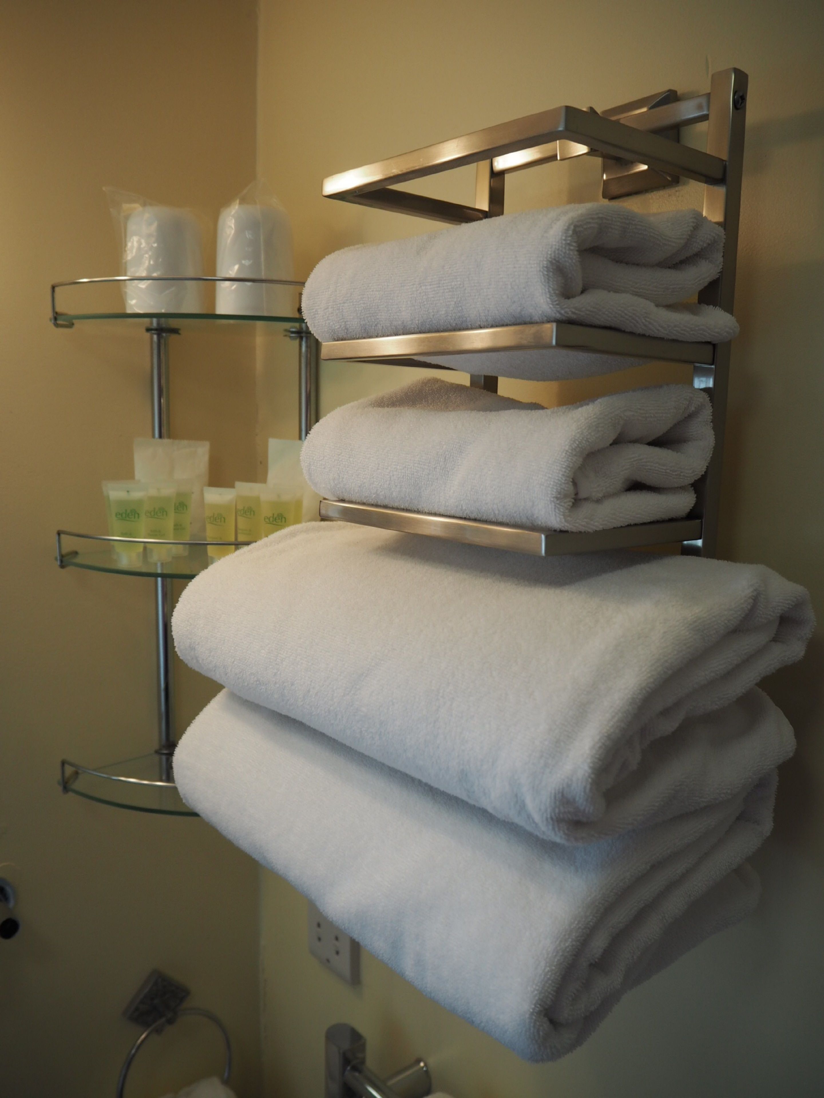 Une salle de bain avec un porte-serviettes sur lequel sont pliés plusieurs serviettes. On y trouve également des étagères en verre où sont rangés les articles de toilette, ainsi qu’une cabine de douche en verre.