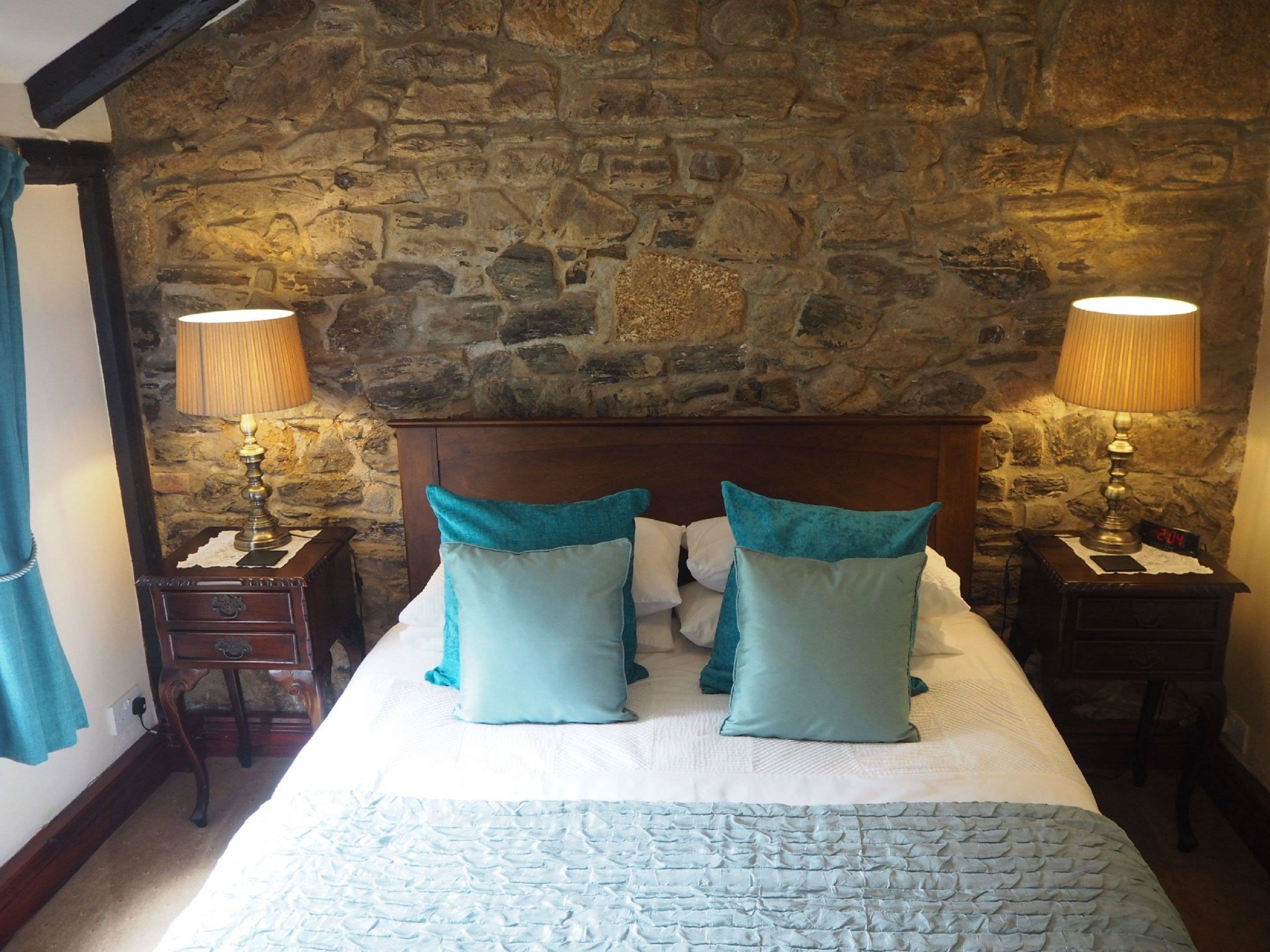 A bedroom with a stone wall, a wooden headboard, two bedside tables with lamps, and a bed with teal pillows.