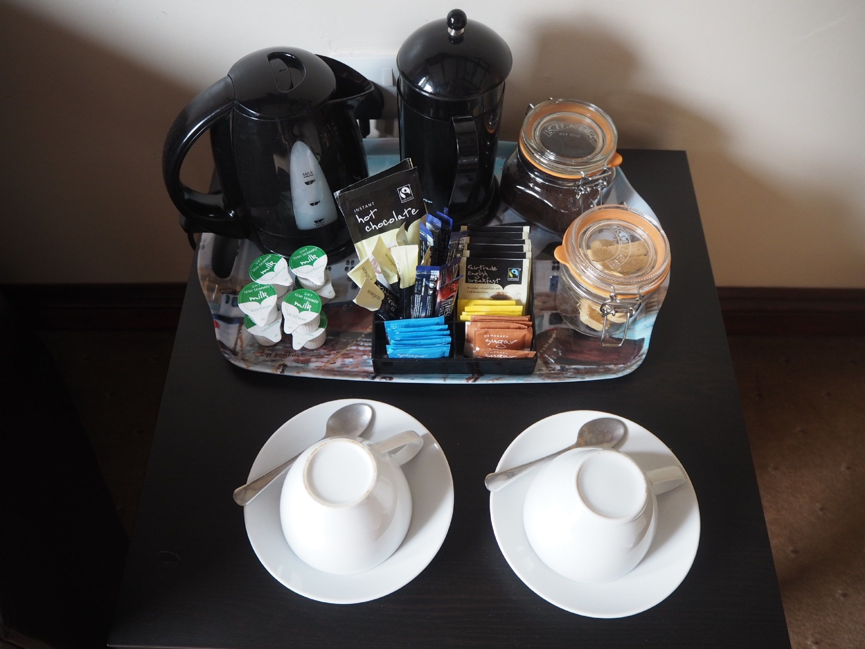 En bakke med kaffemaskine, termokande, et syltetøjsglas og diverse morgenmadsprodukter på et bord.