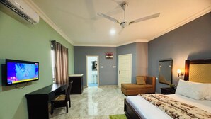 A hotel room with a bed, a desk, a chair, a TV, a ceiling fan, and a bathroom visible through an open door. - Dafika Lodge (Tema)