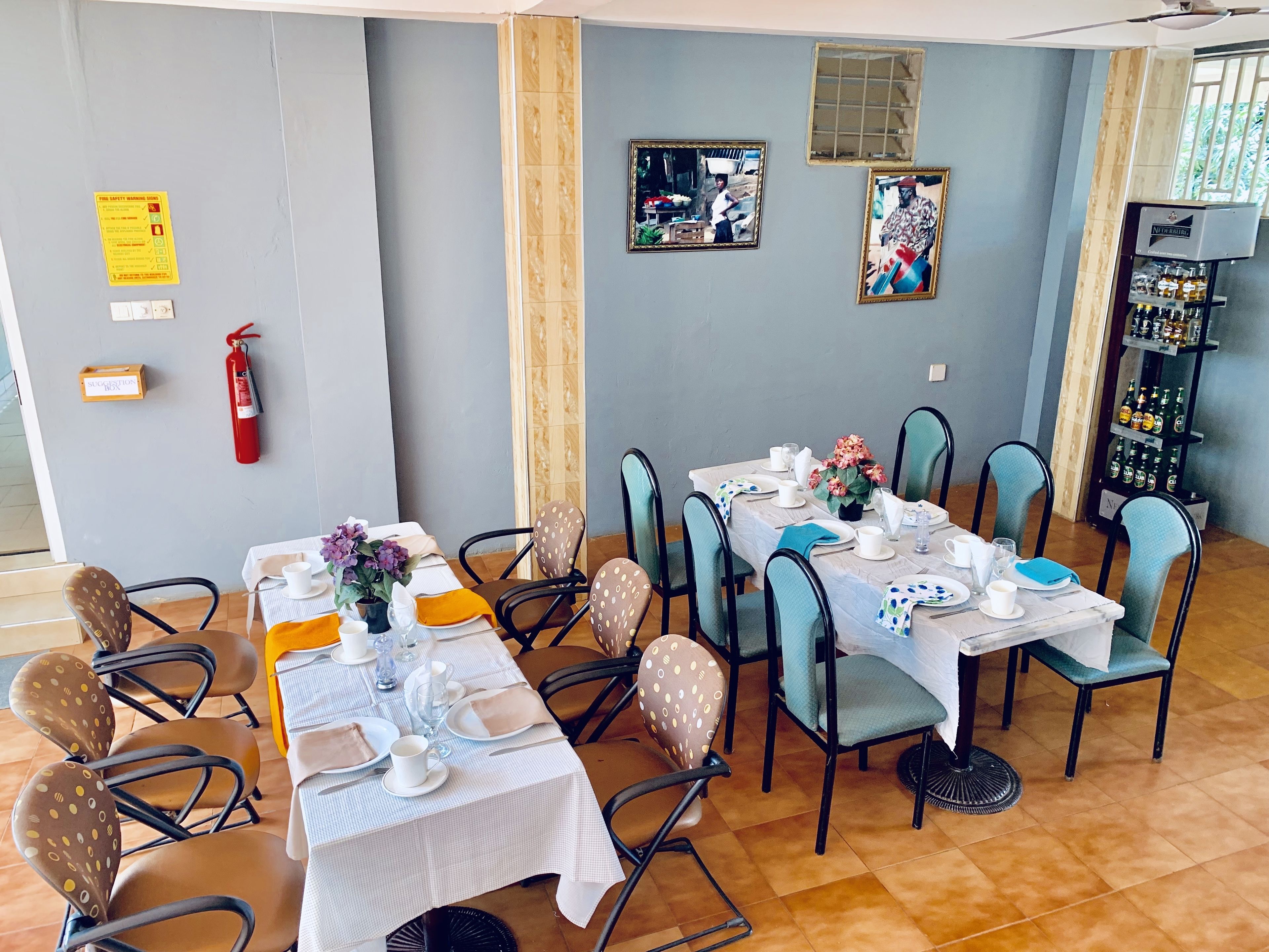 A dining area with tables set for four, chairs with patterned upholstery, a fire extinguisher, a water dispenser, and framed pictures on the wall.