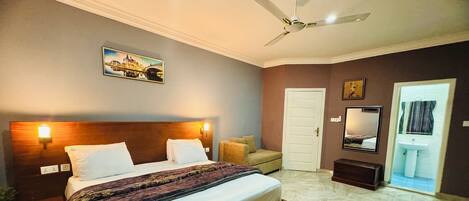 Habitación de hotel con cama, mesita de noche, una silla, ventilador de techo, puerta, espejo y un cuadro colgado en la pared.