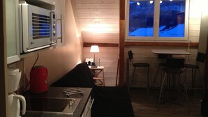 Eine kompakte Küche mit Mikrowelle, Wasserkocher und Herd. Ein kleiner Essbereich mit Tisch und Stühlen ist vorhanden. Durch ein Fenster ist eine verschneite Winterlandschaft zu sehen.