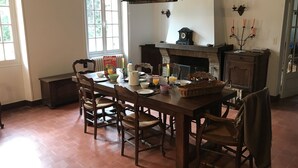 Ein Esszimmer mit einem Holztisch, gedeckt für sechs Personen, umgeben von Stühlen. Eine Kristallleuchter hängt darüber, und im Hintergrund ist ein Kamin zu sehen.