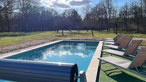 Ein rechteckiger Swimmingpool mit klar blauem Wasser, umgeben von einer Rasenfläche mit Bäumen im Hintergrund. Liegestühle sind am Poolrand aufgereiht.