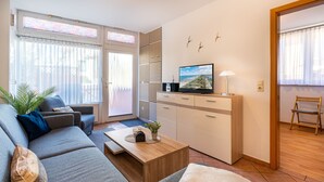 A modern living room with a grey sofa, a wooden coffee table, a TV on a white cabinet, and a view of the outdoors through large windows. - Bungalow Möwe (opposite the “Hus Günter”) (Seebad Ahlbeck, Mecklenburg-Vorpommern)