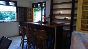Un espace bar doté d’un comptoir en bois, de trois tabourets de bar en osier et d’une étagère où sont exposés des objets décoratifs.