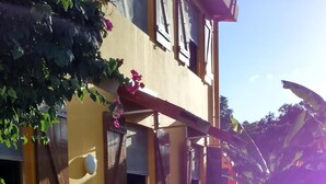 Une terrasse en bois avec une table et des chaises, un pot de fleurs suspendu, et un bâtiment aux murs jaunes et aux volets en bois.