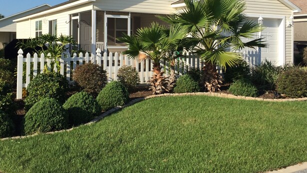A single-story house with a white exterior, a front porch, and a small front yard with a white picket fence and palm trees. - Village of Fernandina - free use of golf cart with liability waiver (The Villages)