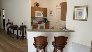 A compact kitchen with wooden cabinets, a microwave, a stove, and a sink. There are two wicker chairs at a small bar area. - Village of Fernandina - free use of golf cart with liability waiver (The Villages)
