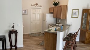 A compact kitchen with wooden cabinets, a refrigerator, and a small dining area with a chair. - Village of Fernandina - free use of golf cart with liability waiver (The Villages)