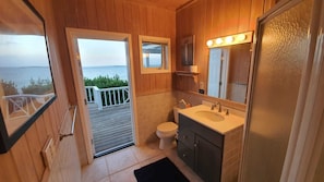 Ein Badezimmer mit Holzausstattung, das eine Toilette, ein Waschbecken und einen Waschtisch, einen Spiegel mit Beleuchtung sowie Meerblick durch eine offene Tür bietet.