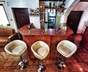 A bar area with a wooden counter, three bar stools, and a refrigerator stocked with beverages.