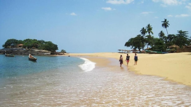 Una spiaggia con acqua cristallina, una riva sabbiosa e alcune persone che passeggiano. Si vedono imbarcazioni in acqua e palme sullo sfondo.