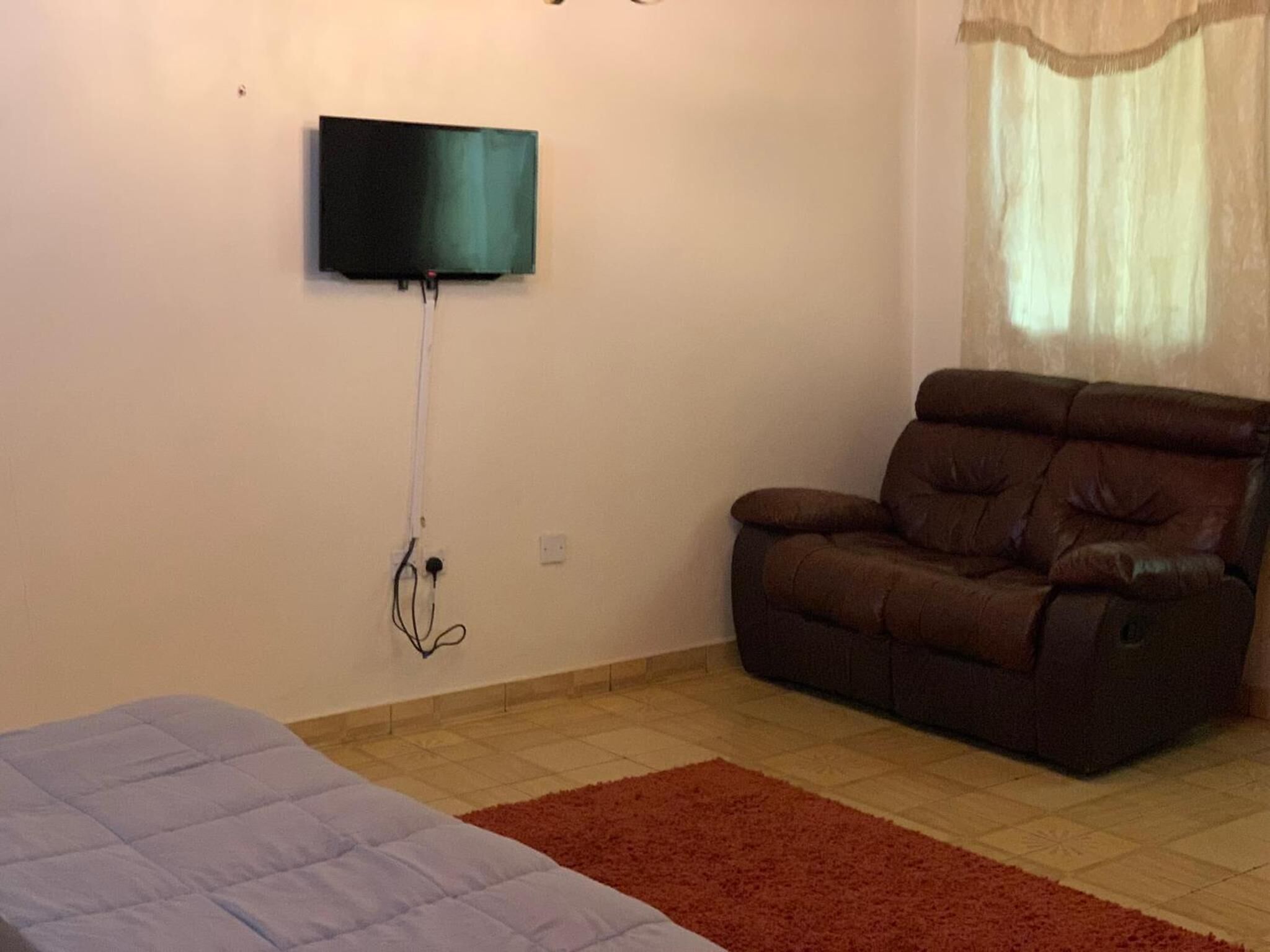 A room with a bed, a brown sofa, a television mounted on the wall, and a window with a sheer curtain.
