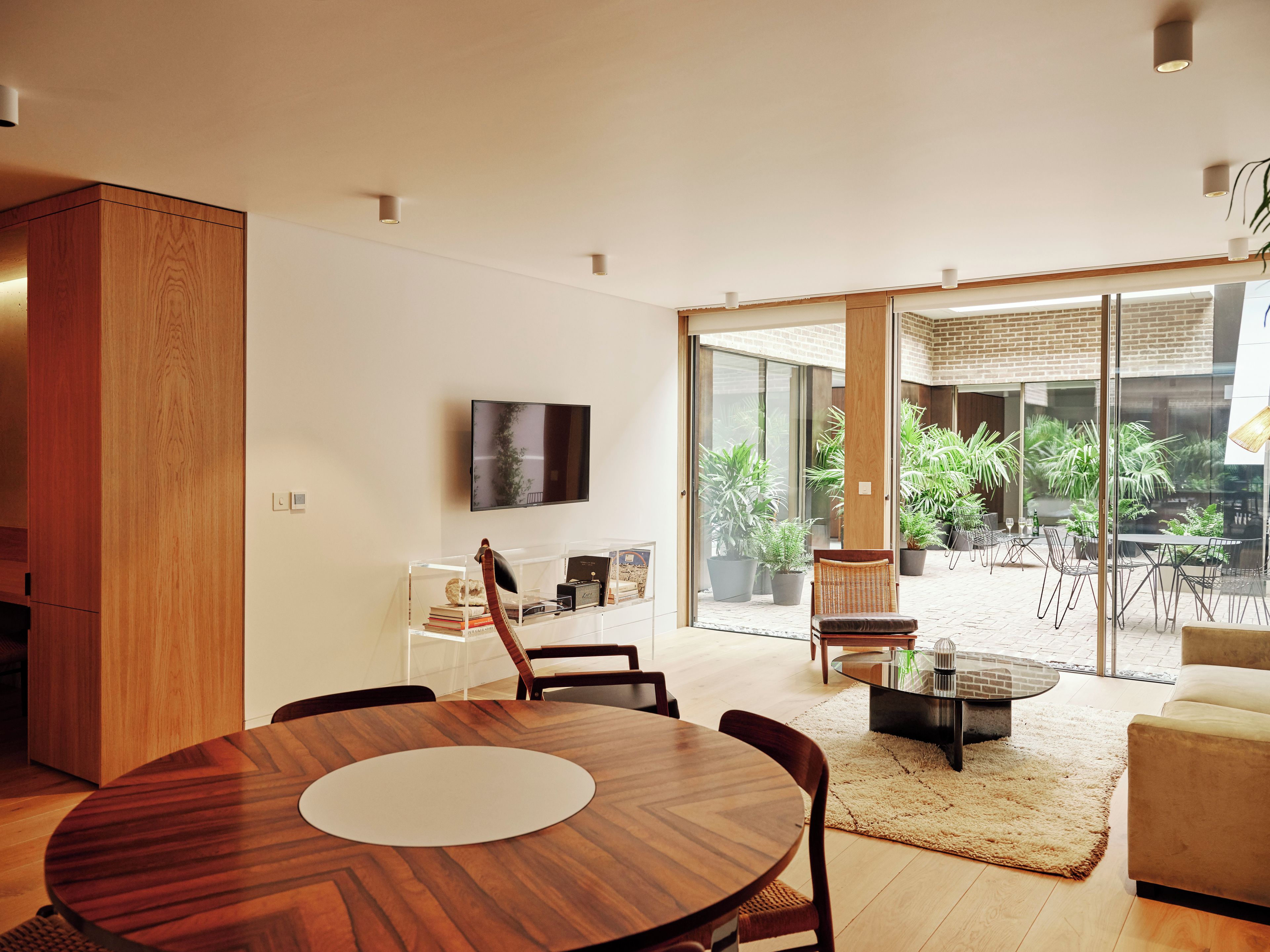 A modern living room with a wooden dining table, a sofa, and a glass door leading to a patio with plants.