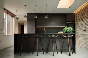 A modern kitchen with dark cabinetry, a central island with bar stools, pendant lighting, and a brick accent wall. - Weymouth Mews (London)