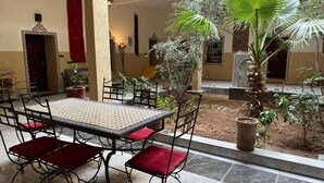 A courtyard with a tiled table and chairs, surrounded by plants and a palm tree. - Chambres d'Hôtes Riad Dar Tazoulte (Agdz)